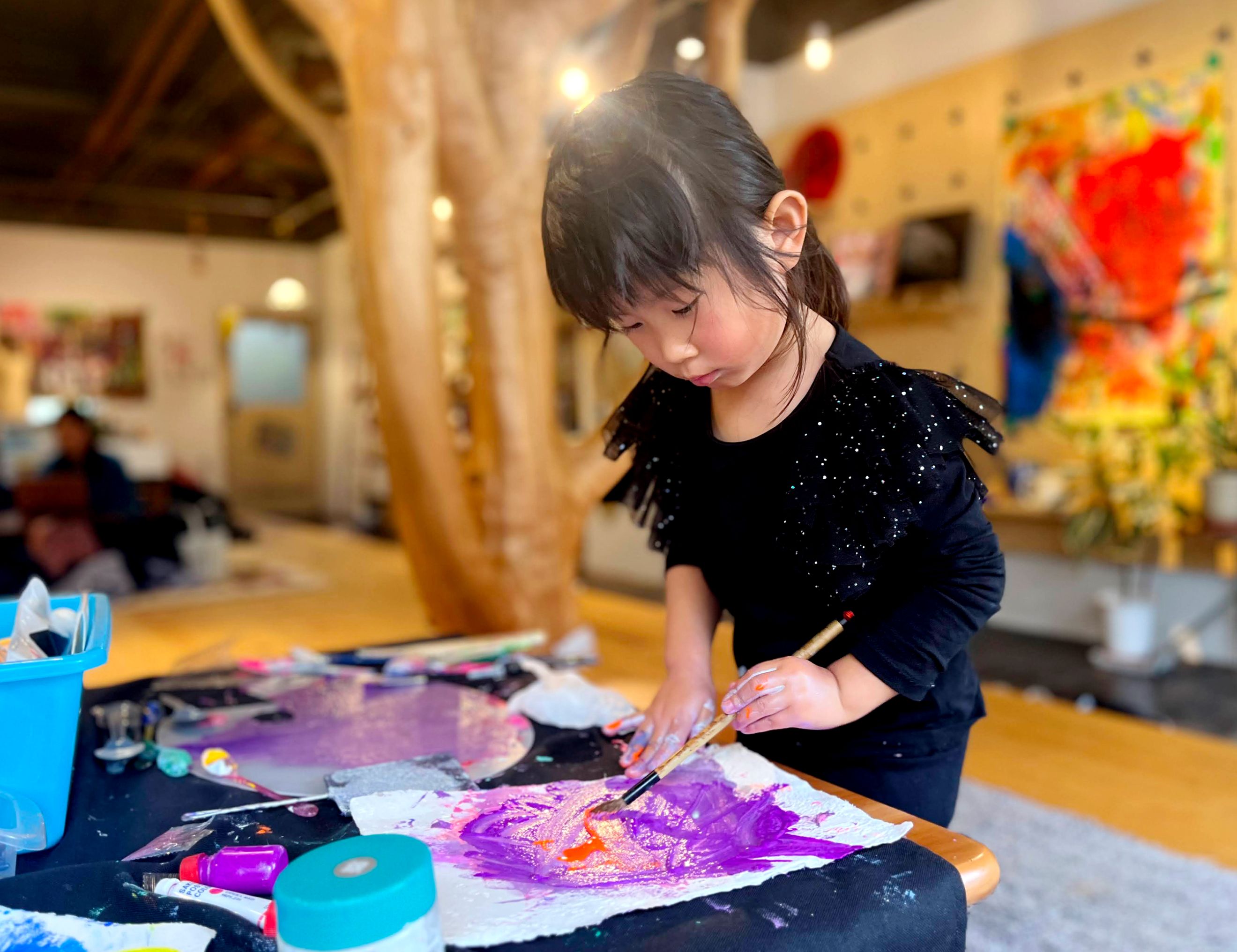 机の上にある大きな紙に紫の絵の具で作品を作っている左利きの女の子