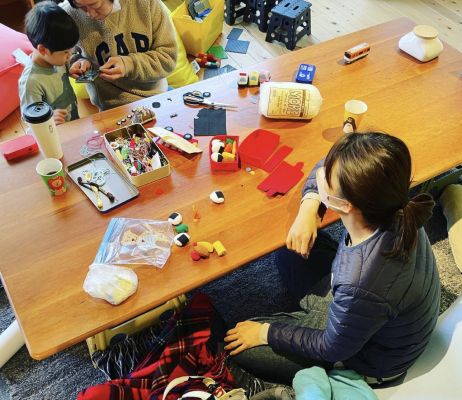 テーブルに座っている子供とお父さんとお母さん。テーブルの上にはさまざまなものが置かれている。子供はお父さんのスマホを覗き込んでいる