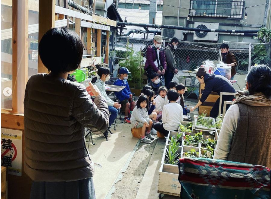 野外で椅子に座っている子供たちとその様子を見ている大人たち