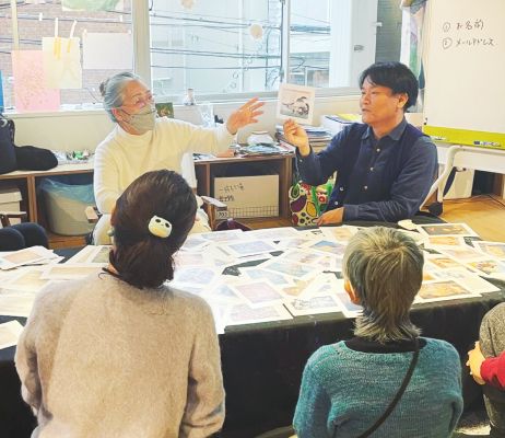 机に座ってパパンダ先生が持っている絵画を見ている3人の女性。机の上にはたくさん絵画が広げられている。
